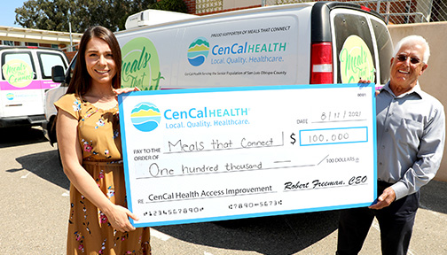 A woman and a man hold a check for $100,000 outside in a parking lot. 